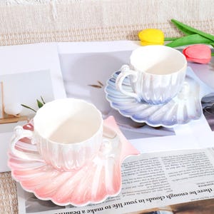 May include: Two ceramic teacups and saucers. The cups and saucers are pearlescent white with a shell design. One saucer is pink and the other is blue. The cups have pearl accents on the handles. The set is on a newspaper.