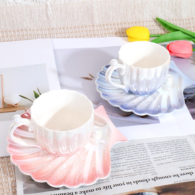 May include: Two ceramic teacups and saucers. The cups and saucers are pearlescent white with a shell design. One saucer is pink and the other is blue. The cups have pearl accents on the handles. The set is on a newspaper.
