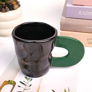 May include: A black ceramic mug with a unique, organic shape and a vibrant green handle. The interior of the mug is black, and the exterior has a glossy finish. The handle is a solid, curved shape, adding a modern touch to the design.