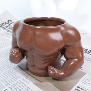 Puede incluir: Una maceta de cerámica marrón con forma de torso musculoso. La maceta tiene una amplia abertura en la parte superior y una escultura detallada del pecho y los brazos. El acabado brillante le da un aspecto pulido, adecuado para la decoración del hogar.