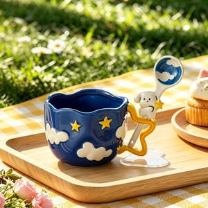 Pode incluir: Uma caneca de cerâmica azul caprichosa com um design de nuvens e estrelas, combinada com uma colher a condizer com um cão e uma estrela. A caneca está numa bandeja de madeira, criando uma estética charmosa e sonhadora.