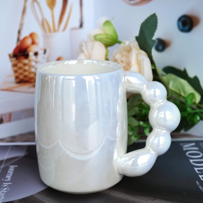 May include: A pearlescent white ceramic mug with a unique handle design. The handle is made of connected spheres. The mug has a glossy finish and is suitable for hot or cold beverages. The mug is a decorative item.