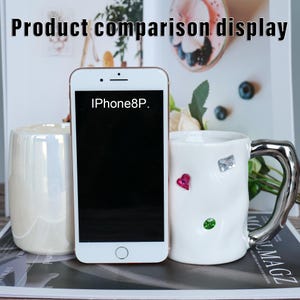 May include: A white iPhone 8P is displayed between a pearlescent ceramic mug and a white mug with a silver handle. The white mug has decorative gems and a heart. The text "Product comparison display" is at the top.