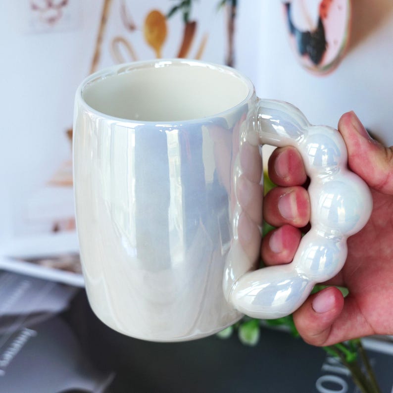 May include: Iridescent white ceramic mug with a unique, bubble-shaped handle. The mug has a pearlescent finish, reflecting light with a subtle sheen. The interior is a clean, bright white. A stylish and modern coffee or tea cup.