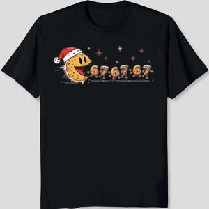 May include: Black t-shirt featuring a cartoon design of a smiling, yellow, Pac-Man-like character wearing a Santa hat. The character is followed by the numbers "67-67-67-67" in a similar style. Red and gold stars are scattered above the characters.