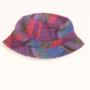 May include: A colorful plaid bucket hat in shades of purple, red, blue, and green. The hat features a classic bucket shape with a slightly downward-sloping brim. The fabric appears to be a woven material.