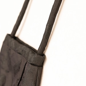May include: A close-up of a black shoulder bag with a textured pattern. The bag has a rectangular shape with a strap and is made of a dark fabric. The strap has a decorative design.