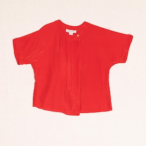 May include: A bright red short-sleeved blouse with a round neckline and a pleated front. The blouse has a single button closure and the brand name "ARGENTI" is visible. The top is made of a lightweight fabric.