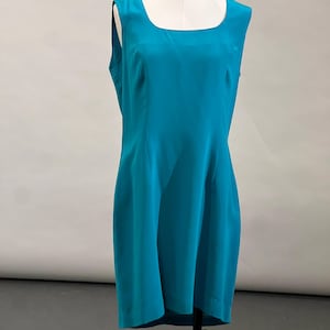 May include: A sleeveless, knee-length dress in a solid, vibrant teal color. The garment features a square neckline, wide shoulder straps, and a straight, tailored silhouette that follows the natural lines of the torso down to the hem.