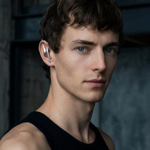 Op de afbeelding: Een zilveren bliksemschicht oormanchet wordt op het linkeroor gedragen. De oormanchet is een uniek sieraad. De persoon op de foto draagt een zwarte tanktop. De achtergrond is een gedempt grijs.
