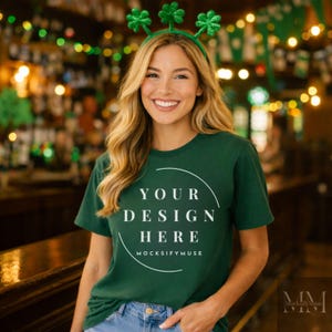 May include: A dark green t-shirt with the text "YOUR DESIGN HERE" in a white semi-circle. The model is wearing a green shamrock headband. The background is a blurred bar scene with warm lighting.