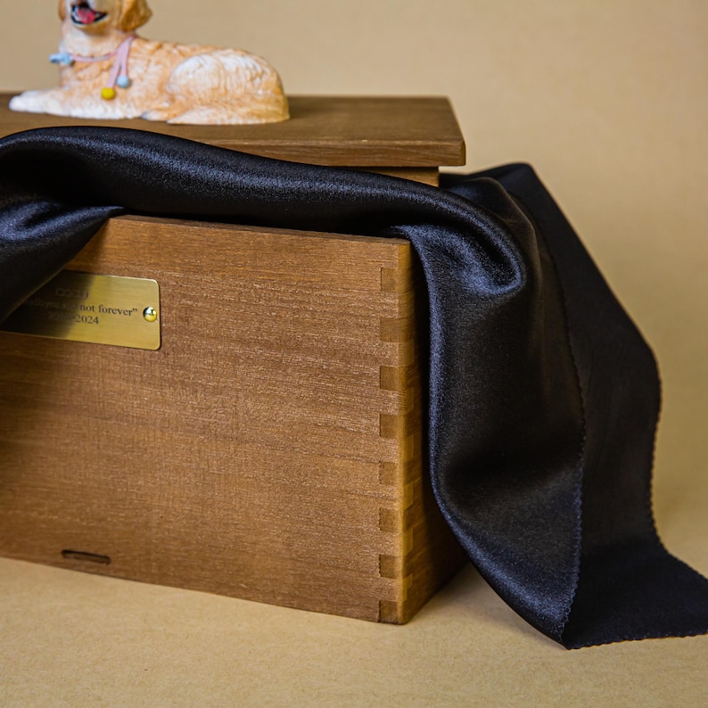 May include: A wooden pet urn with a decorative golden retriever figurine on top. A black satin cloth drapes over the urn. The urn has a brass plaque with text. The wooden box is constructed with dovetail joints.