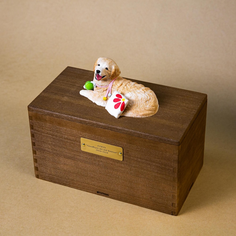 May include: A wooden pet urn with a decorative golden retriever figurine on top. The figurine is lying down with a green ball and a small pillow with a red flower. A gold plaque on the front reads "Coco, Goodbye is not forever 2016-2024."