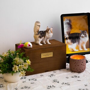 May include: A wooden pet urn with a decorative cat figurine on top. The cat is brown, white, and black. A framed photo of a cat is to the right. A small mouse toy and flowers are in the foreground. The urn has a gold plaque.