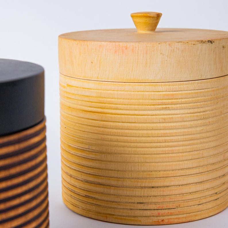 May include: Two wooden storage containers. One is a light-colored cylinder with a lid and a small knob. The other is a dark cylinder with a black lid and brown and orange stripes. The containers are on a white background.