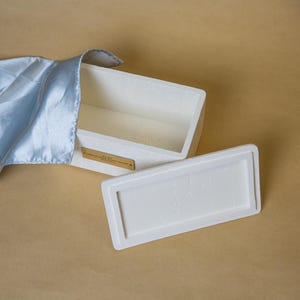 May include: A white rectangular box with a matching lid, open to reveal the interior. A light blue satin cloth is draped over the side. A gold-colored plaque is affixed to the front of the box. The box is on a tan surface.