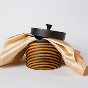 May include: A round wooden box with a lid, featuring a dark brown top and a body with horizontal stripes of brown and yellow. A gold-colored cloth is draped over the box. The box has a small, dark knob on the lid.