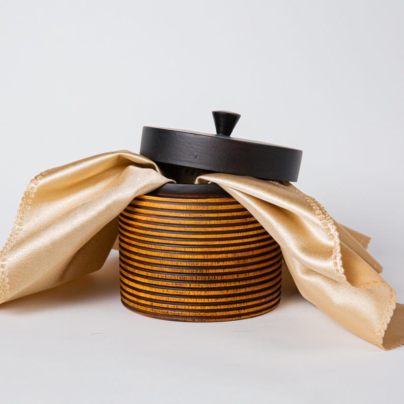 May include: A round wooden box with a lid, featuring a dark brown top and a body with horizontal stripes of brown and yellow. A gold-colored cloth is draped over the box. The box has a small, dark knob on the lid.