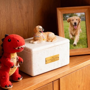 May include: A cream-colored pet urn with a golden retriever figurine on top, a framed photo of a golden retriever, and a red dinosaur plush toy. The urn has a gold plaque with text.
