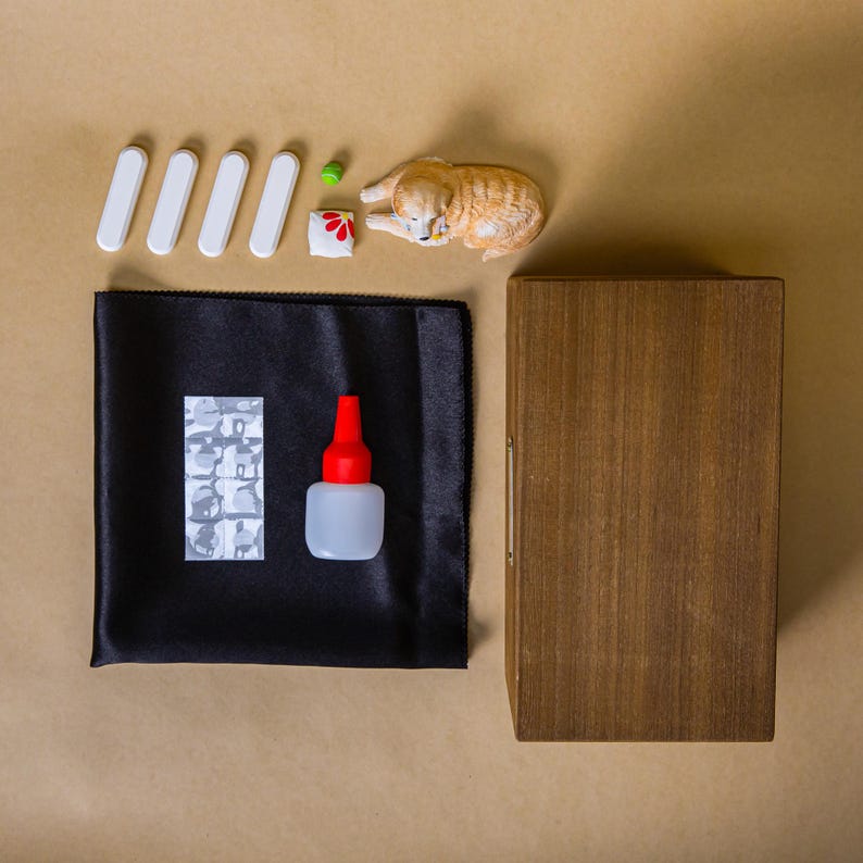 May include: An overhead shot features a collection of items on a tan surface. There are five white, rectangular pieces, a small green ball, a miniature cat figurine, and a black cloth with a patterned patch. A small bottle with a red cap and a wooden box are also present.