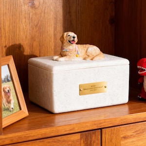 May include: A cream-colored pet urn with a sculpted golden retriever figurine on top. The urn has a gold plaque with the name "Ruby" and the inscription "Always loved, forever missed" and the years 2020-2023.