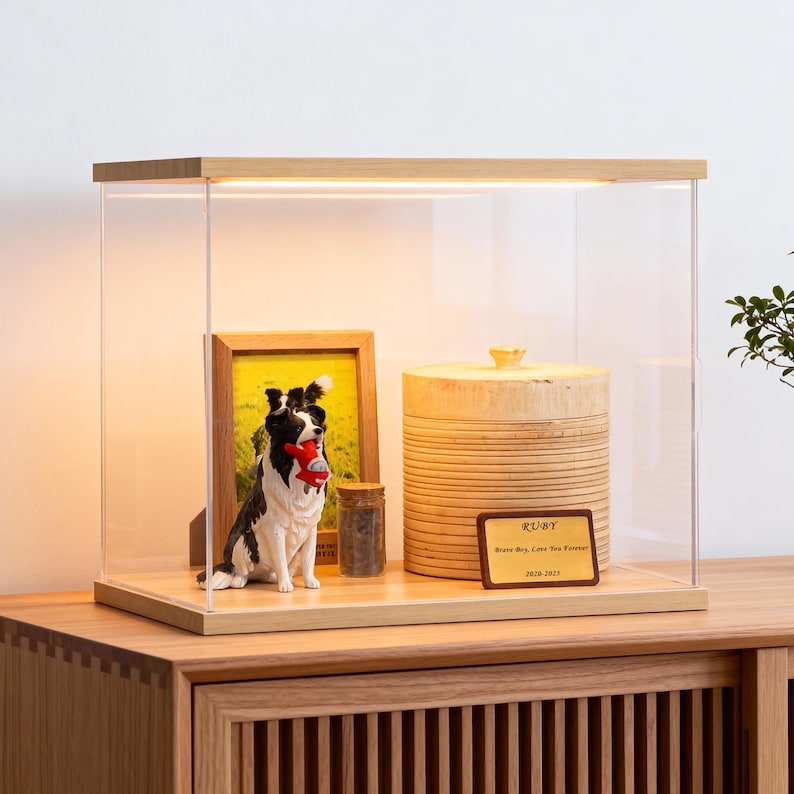 May include: A wooden display case with a clear acrylic front houses a framed photo, a dog figurine holding a red toy, a small jar, and a wooden container with a lid. A plaque reads "RUBY" and a message.