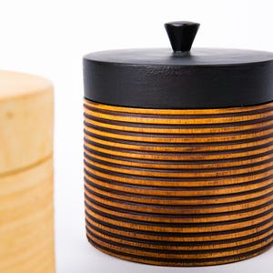 May include: A cylindrical wooden container with a black lid and a small, conical handle. The body of the container is made of horizontal, alternating bands of light and dark brown wood. A second, lighter-colored wooden container is partially visible in the background.