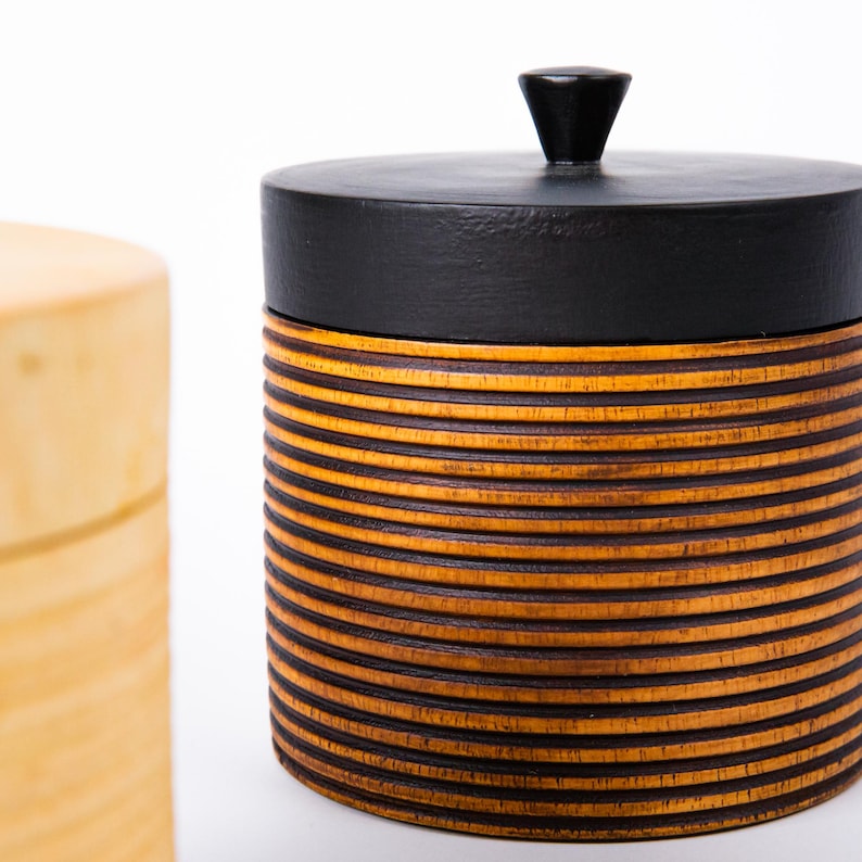 May include: A cylindrical wooden container with a black lid and a small, conical handle. The body of the container is made of horizontal, alternating bands of light and dark brown wood. A second, lighter-colored wooden container is partially visible in the background.