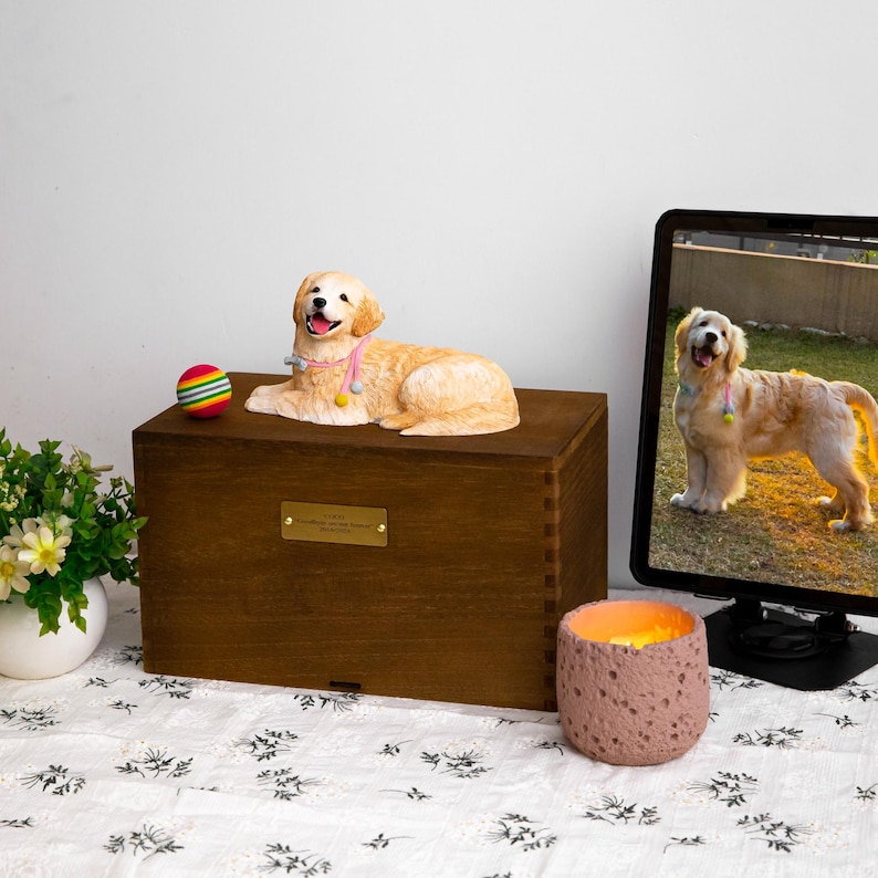 May include: A wooden pet urn with a golden retriever figurine on top. The dog is wearing a pink collar with colorful pom-poms. A colorful ball sits next to the dog. A lit candle and a computer monitor with a dog image are also in the image.