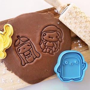 May include: A wooden rolling pin with star patterns, alongside yellow, blue, and brown cookie cutters in the shapes of a cartoon dog, penguin, and girl. The cookie cutters are on a wooden cutting board with rolled-out dough.