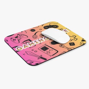 May include: A rectangular mousepad with a gradient of pink, orange, and yellow. The mousepad features a pattern of gaming-related doodles, including game controllers, headphones, and a sword. The word "GAMING" is prominently displayed.