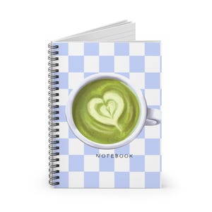 May include: Spiral-bound notebook with a light blue and white chequered pattern. The cover features a cup of matcha latte with a heart-shaped design in the foam. The word "NOTEBOOK" is printed at the bottom.