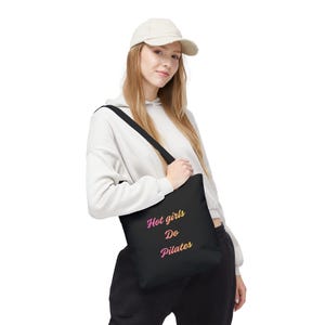 Puede incluir: Bolso tote negro con el texto "Hot girls Do Pilates" en degradado rosa y amarillo. El bolso tiene una correa negra para el hombro. La persona de la imagen lleva una sudadera con capucha blanca y una gorra de béisbol color crema.