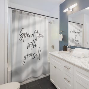 May include: A white shower curtain with the words "Spend time with yourself" in black script. The bathroom features white cabinets, a marble countertop, and a dark grey tiled floor. A mirror reflects the shower curtain.