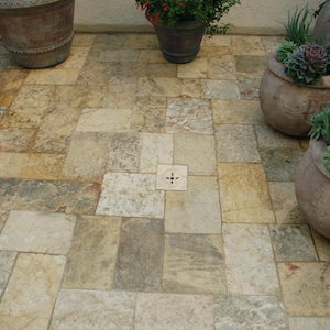 Op de afbeelding: Een stenen terras met rechthoekige en vierkante tegels in beige, grijs en bruin. Verschillende grote, verweerde terracotta potten met planten zijn zichtbaar, waaronder vetplanten en bloeiende planten.