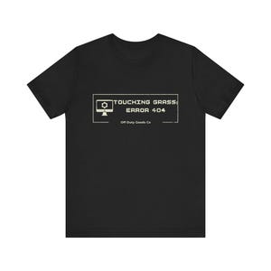 May include: Black t-shirt with a white graphic that reads "TOUCHING GRASS: ERROR 404" above the text "Off Duty Goods Co." The graphic features a computer monitor icon. The shirt is made of a soft material.