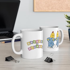 May include: Two white ceramic mugs with the words "BANANAS in PYJAMAS" in colorful block letters. One mug features two cartoon bananas in blue and white striped pajamas. The mugs are on a light-colored surface.