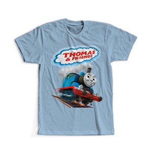 May include: Light blue t-shirt featuring a graphic of Thomas the Tank Engine from Thomas & Friends. The image shows Thomas in motion with a red and blue design, and the text "THOMAS & FRIENDS" in a cloud-shaped frame.