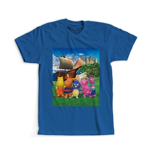 Könnte beinhalten: Ein blaues T-Shirt mit einem farbenfrohen Aufdruck von Zeichentrickfiguren vor einem Piratenschiff, einer Burg und einer Berglandschaft. Die Figuren sind in verschiedenen Farben gehalten, darunter Gelb, Orange, Blau und Rosa. Das Shirt ist aus weichem Material.
