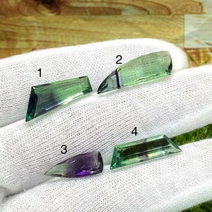 May include: Four faceted gemstones labeled 1, 2, 3, and 4. Gemstone 1 is green with a purple band. Gemstone 2 is light green with a dark purple stripe. Gemstone 3 is deep purple and green. Gemstone 4 is green with a thin purple line, all on white fabric.