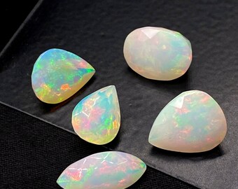May include: Five opals with iridescent color flashes are displayed against a dark background. The gemstones vary in shape, including oval, teardrop, and rounded forms. Faceted surfaces catch the light, showcasing a spectrum of colors.