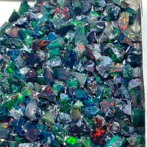 Op de afbeelding: Een verzameling ruwe, ongeslepen opalen in verschillende tinten groen, blauw en zwart. De opalen hebben een natuurlijke, onregelmatige vorm en vertonen flitsen van iriserende kleur. De edelstenen liggen in een metalen bakje.