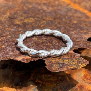 Könnte beinhalten: Ein Ring aus poliertem Silber mit einem durchgehenden gedrehten Seildesign. Das Band ist aus einem einzigen Stück Metall gefertigt und weist ein sich wiederholendes Spiralmuster auf, das eine texturierte, abgerundete Optik erzeugt.