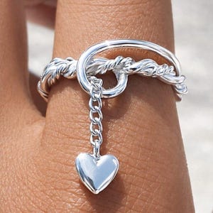 May include: A silver ring with a heart charm. The ring has a twisted rope design and a dangling heart pendant attached by a small chain. The heart is smooth and polished, reflecting light. The ring is worn on a finger.