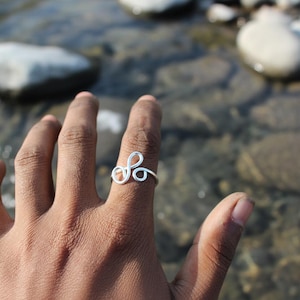 Op de afbeelding: Een zilverkleurige draadring met een lusontwerp wordt op een vinger gedragen. Het ontwerp van de ring heeft een reeks rondingen en cirkels. De achtergrond is wazig en toont een natuurlijke buitenomgeving met water en rotsen.