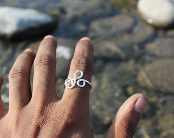 Peut inclure: Une bague en fil d'argent avec un motif en boucle est portée au doigt. Le design de la bague présente une série de courbes et de cercles. L'arrière-plan est flou, montrant un cadre extérieur naturel avec de l'eau et des rochers.
