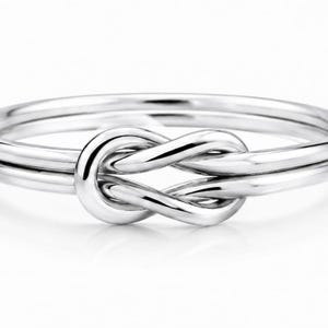 Peut inclure: Une bague argentée polie présentant un motif de nœud infini central. L'anneau est composé de deux fils parallèles qui convergent vers un nœud lisse et symétrique à l'avant, créant une surface métallique continue, élégante et réfléchissante.