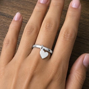 May include: A silver ring with a dangling heart charm. The heart is a solid, polished shape, suspended from a small loop on the ring band. The ring is shown on a hand with a natural skin tone.