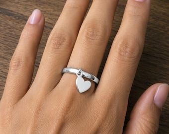 Può includere: Un anello d'argento con un ciondolo a forma di cuore. Il cuore è sospeso da un piccolo anello sulla fascia dell'anello. L'anello è mostrato su una mano con un incarnato naturale, su sfondo di legno.