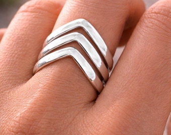 Può includere: Primo piano di un anello d'argento con un design a forma di freccia. L'anello ha tre fasce sovrapposte, ognuna a forma di V. L'anello è indossato su un dito e il metallo ha una superficie lucida e riflettente.